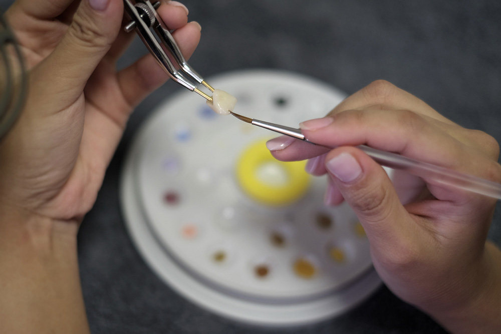 Foto: Zahntechniker wendet Keramikmaterial auf die Krone mit Pinzette und Pinsel auf Zahnpalette Hintergrund. Dentaltechnik Nahaufnahme